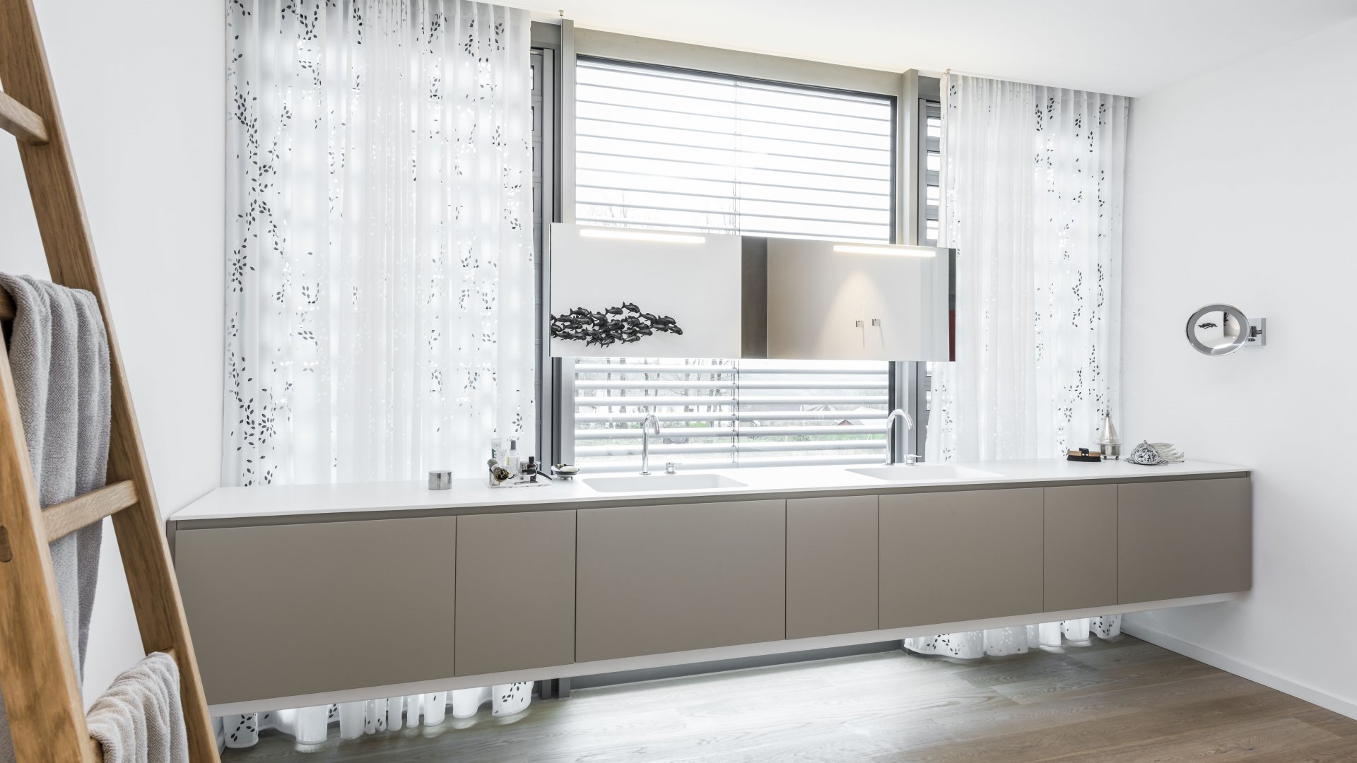 Waschtischanlage aus mattweißem Corian, Handtuchleiter aus Holz, Waschtisch vor Fenster, Vorhang, hängender Spiegel beleuchtet